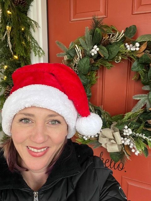 A woman wearing a santa hat is smiling in front of a christmas wreath.