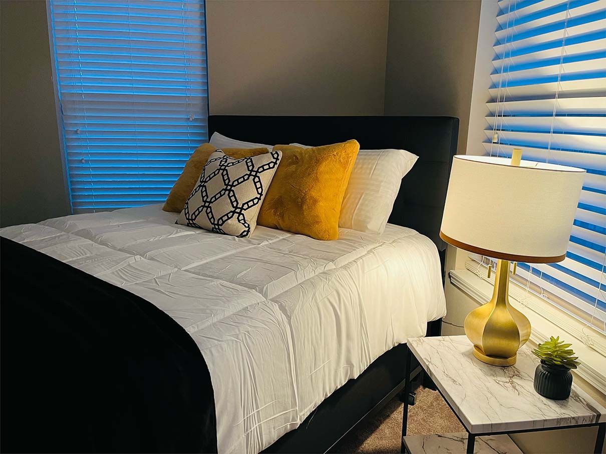 A bedroom with a bed , nightstand , lamp and window with blinds.