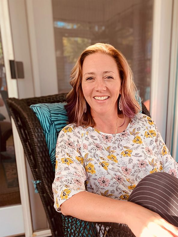 A woman is sitting in a wicker chair on a porch and smiling.