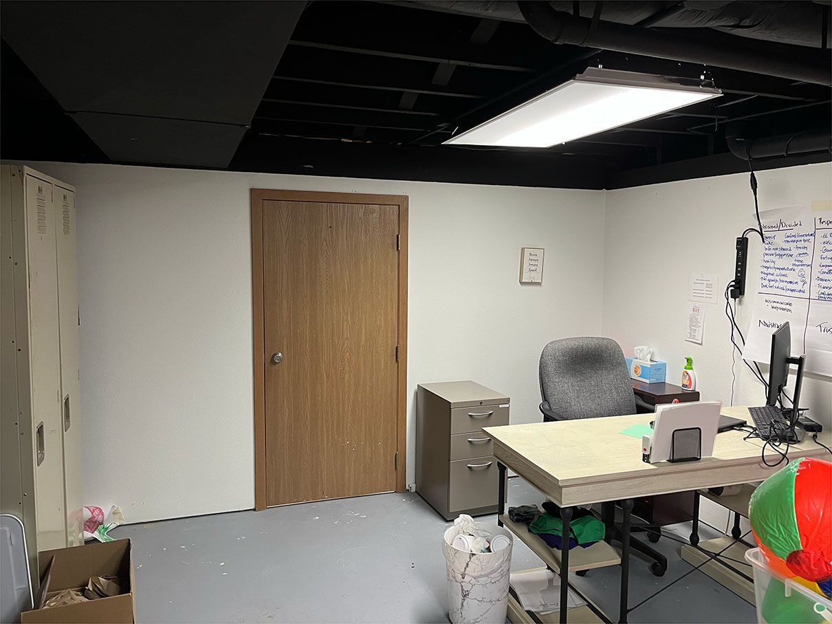 A basement office with a desk , chair , lockers and a computer.