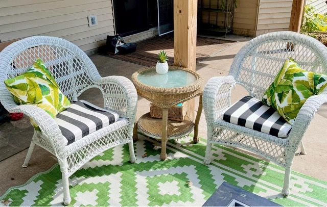 Two wicker chairs and a table on a patio with a green rug.