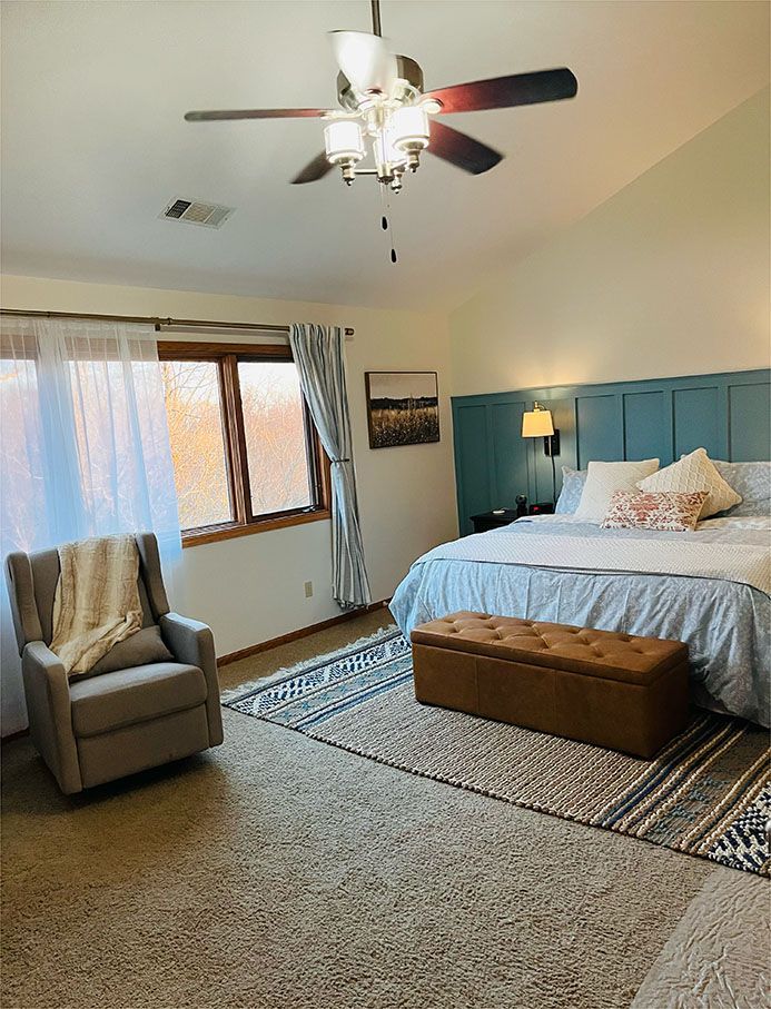 A bedroom with a bed , chair , ottoman and ceiling fan.