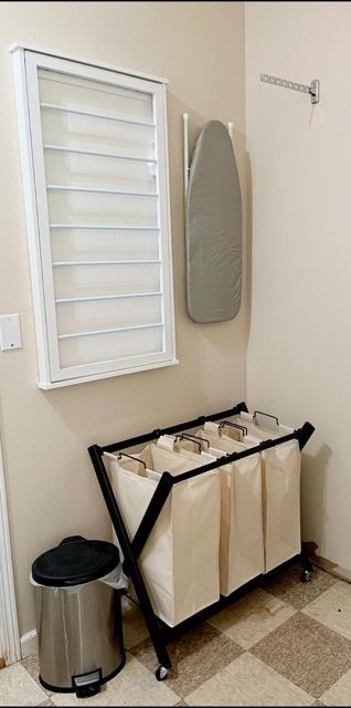 A laundry room with a laundry basket , ironing board , and trash can.