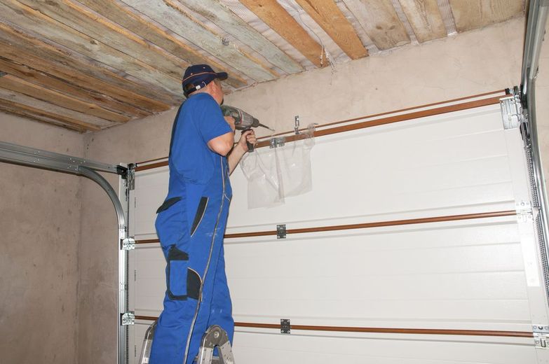 Person in blue coveralls using a drill to install garage door components. Inside, ceiling beams visible.