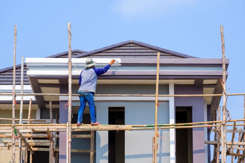 Construction worker painting the exterior of a house from scaffolding. Bright blue sky overhead.
