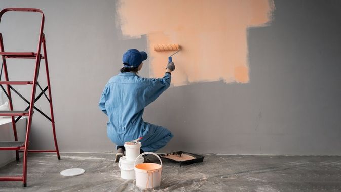 Person painting an orange square on a gray wall with a paint roller; ladder and paint cans nearby.