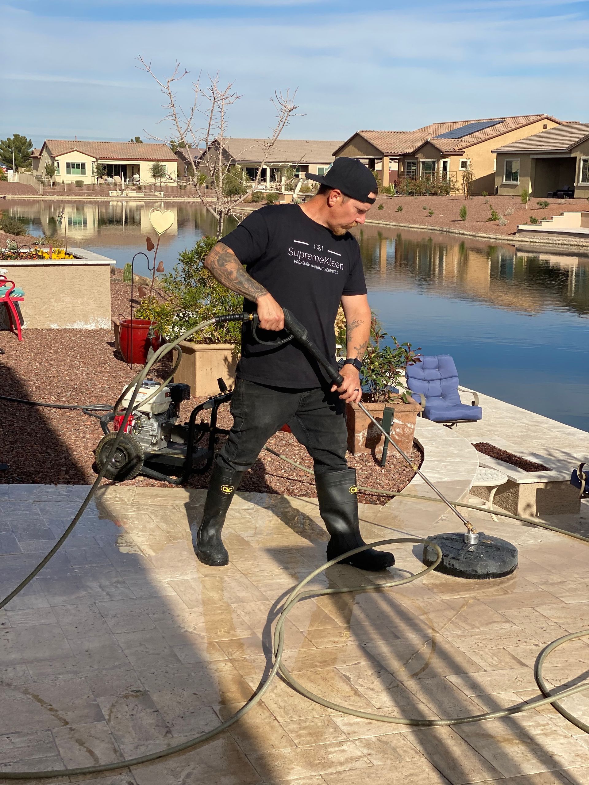 Man using a pressure washer to clean a patio near a pond; houses in the background.