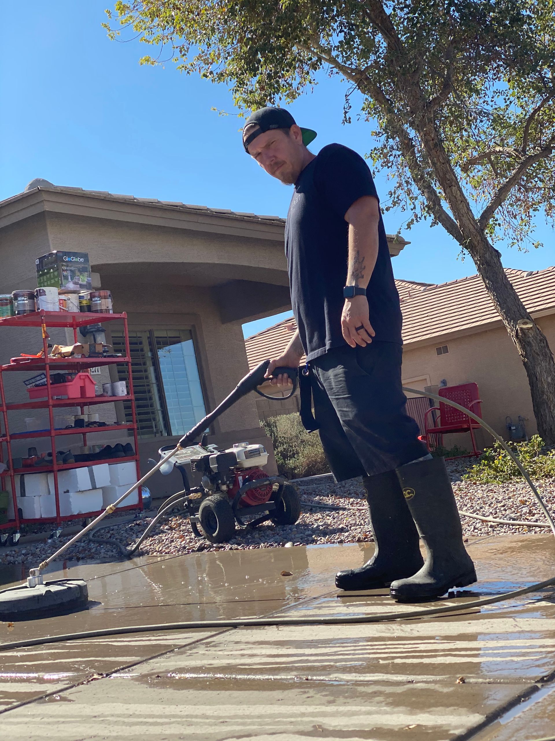 Man pressure washing a driveway wearing a black shirt, cap, and boots on a sunny day.