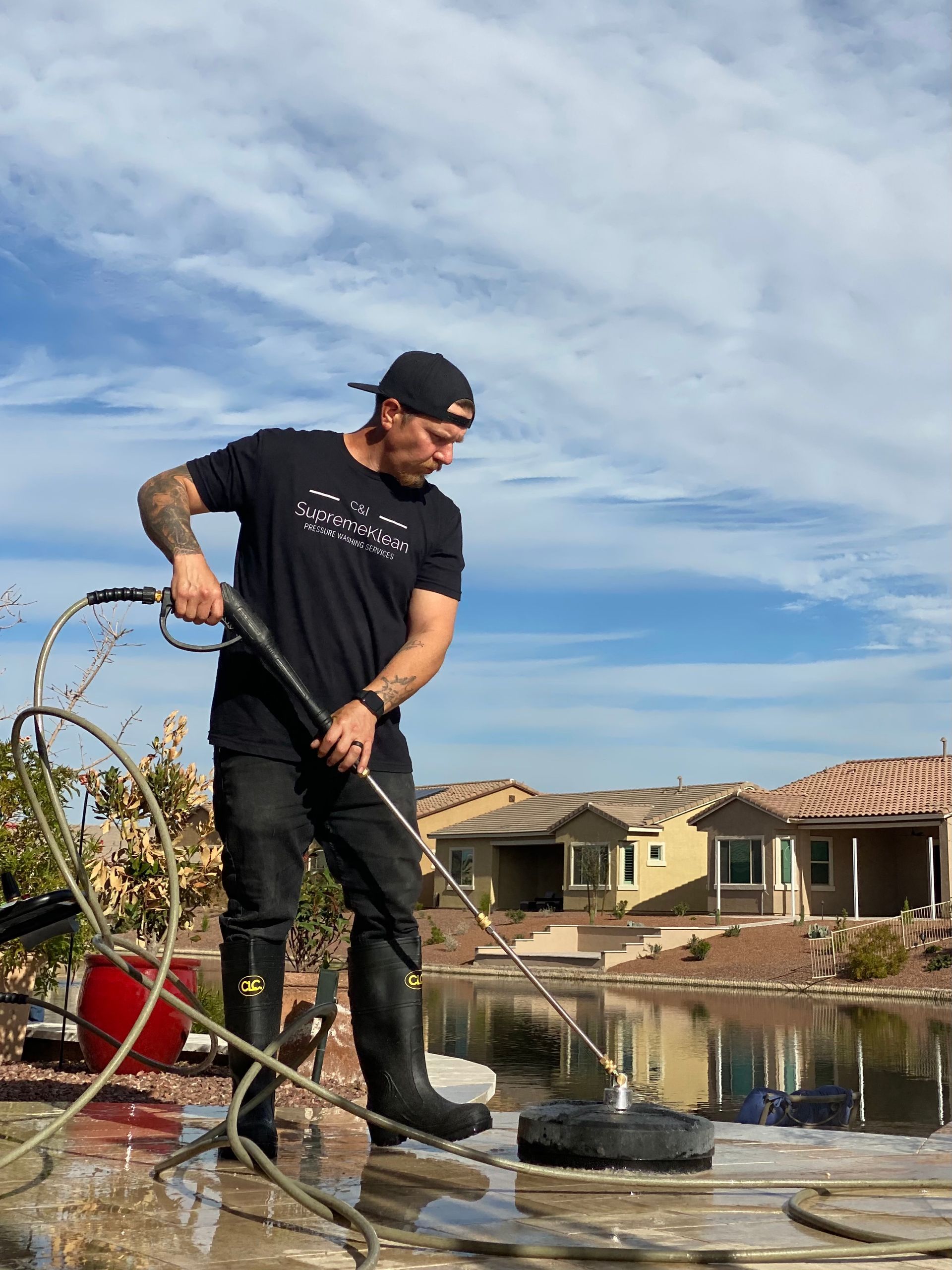 Man using a power washer near a pond. Wearing black clothing, cap and boots.