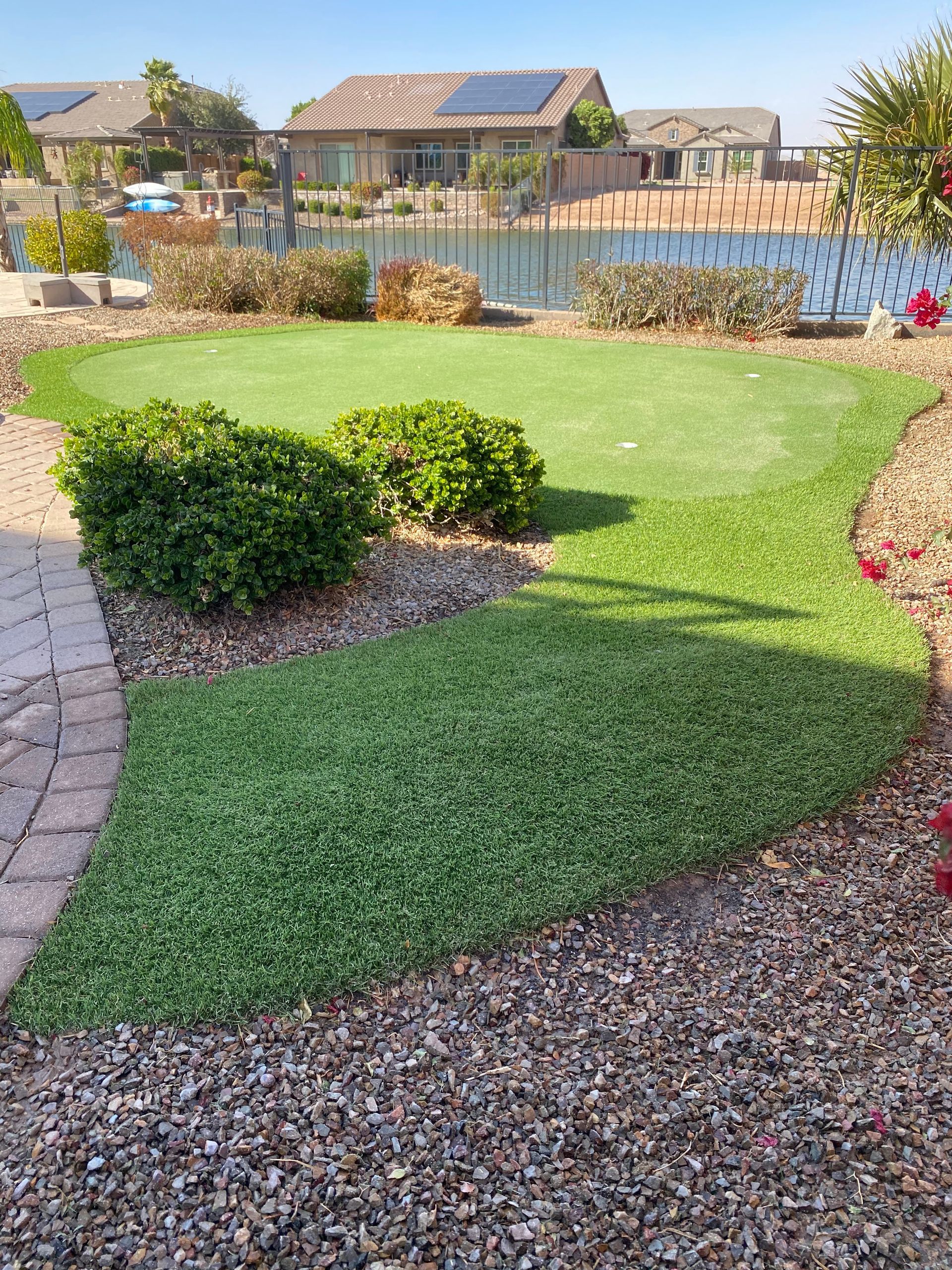 Green lawn and bushes bordering a pebbled area, with houses and a lake in the background.