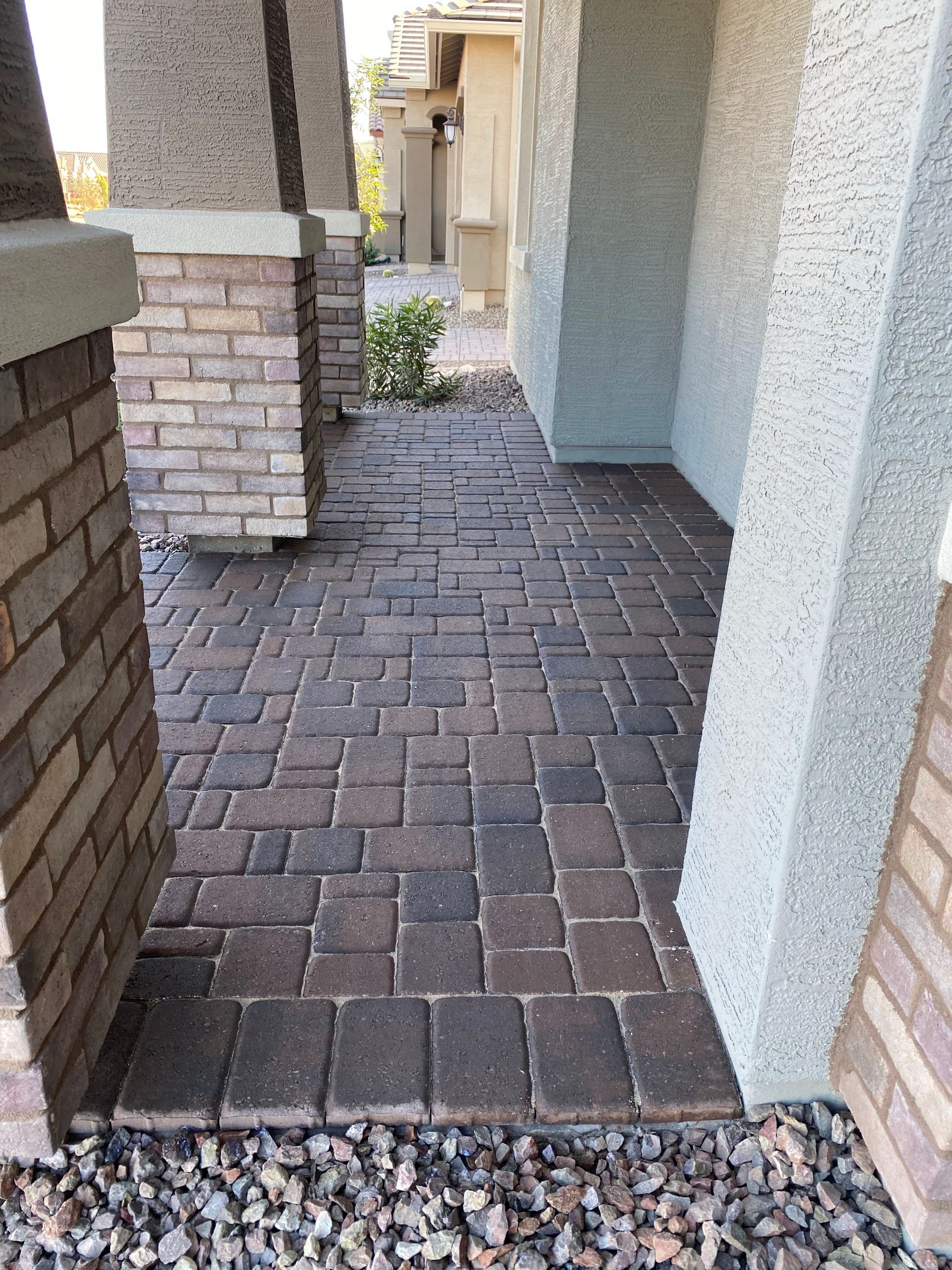 Brick pathway leading to a house entrance, flanked by columns and a gravel border.