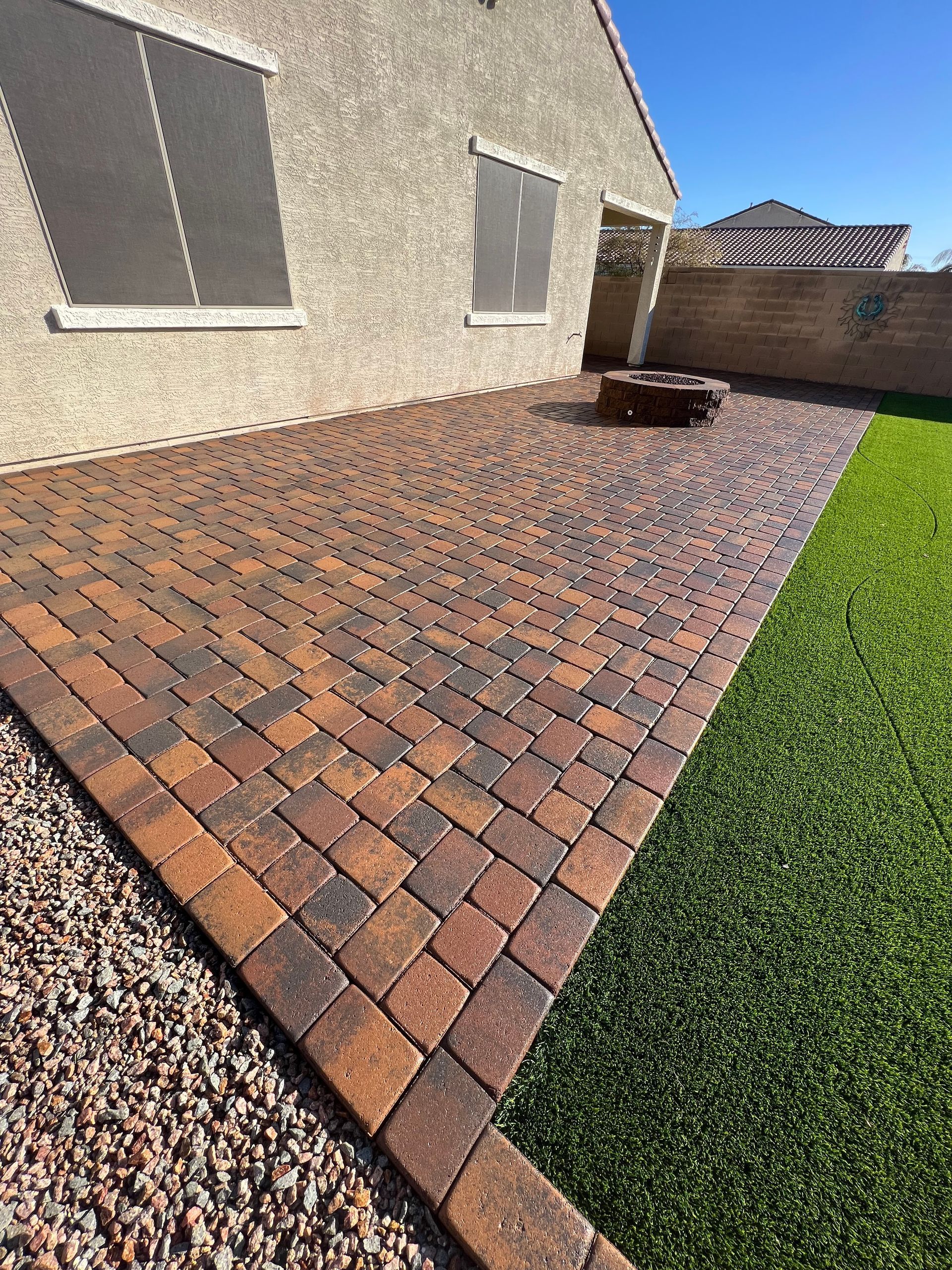 Brick patio next to artificial grass, small fire pit, and tan stucco home exterior on a sunny day.