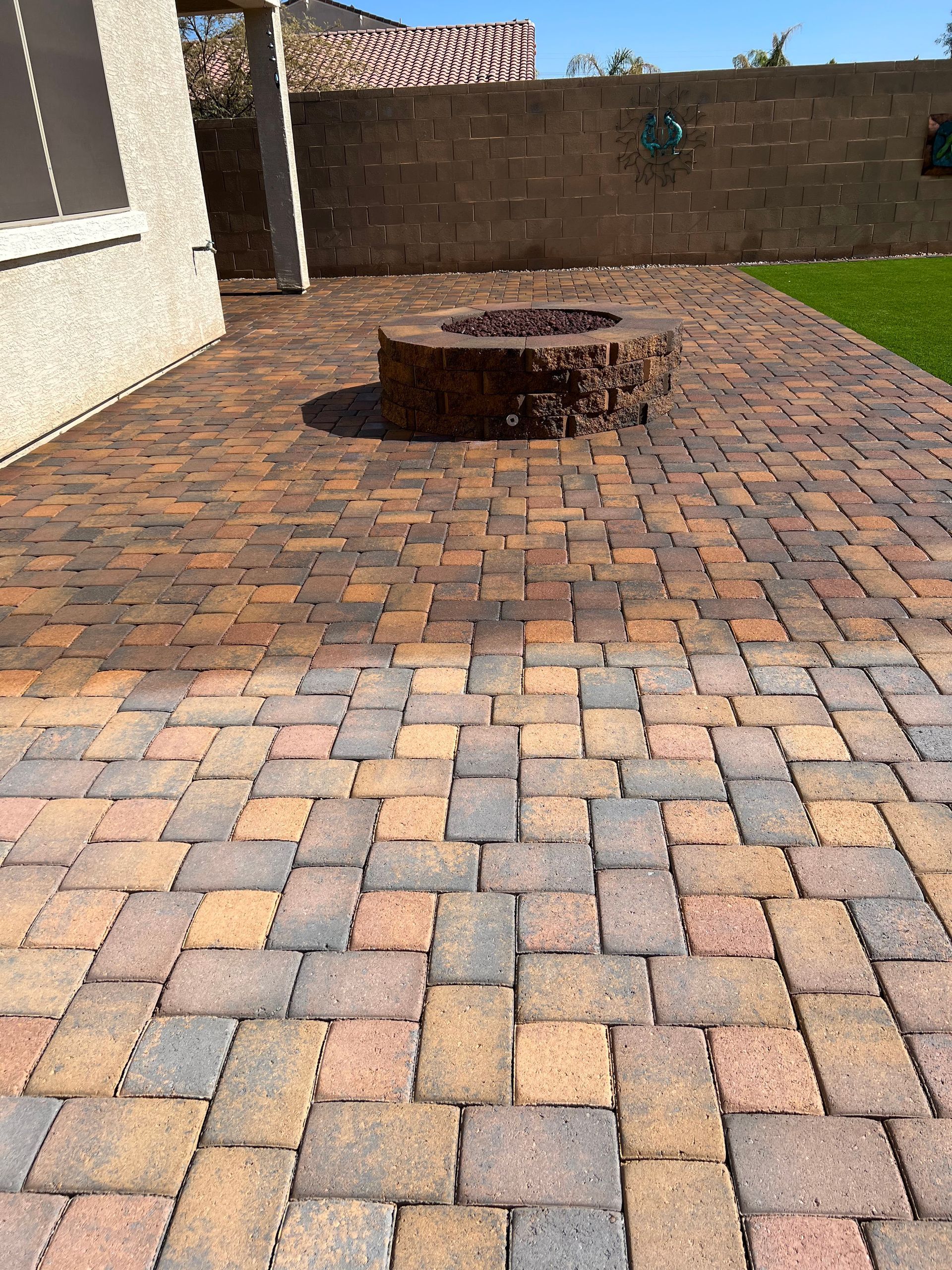 Brick patio with fire pit, bordered by grass and a wall, next to a building.