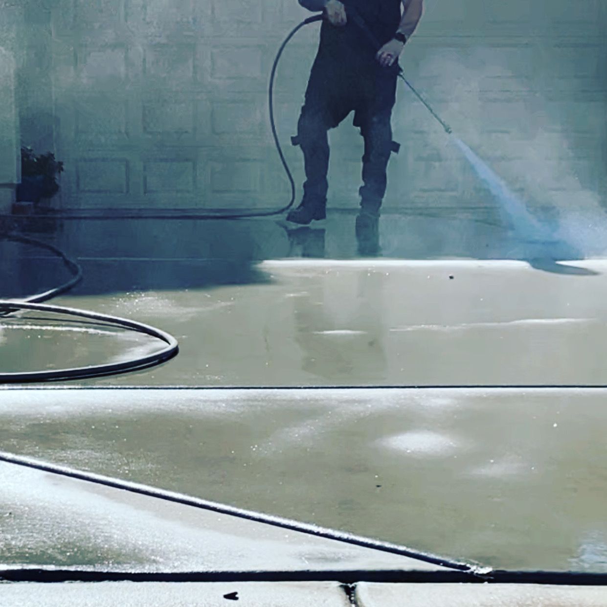 Person using a pressure washer on a concrete driveway, spraying water towards the camera.