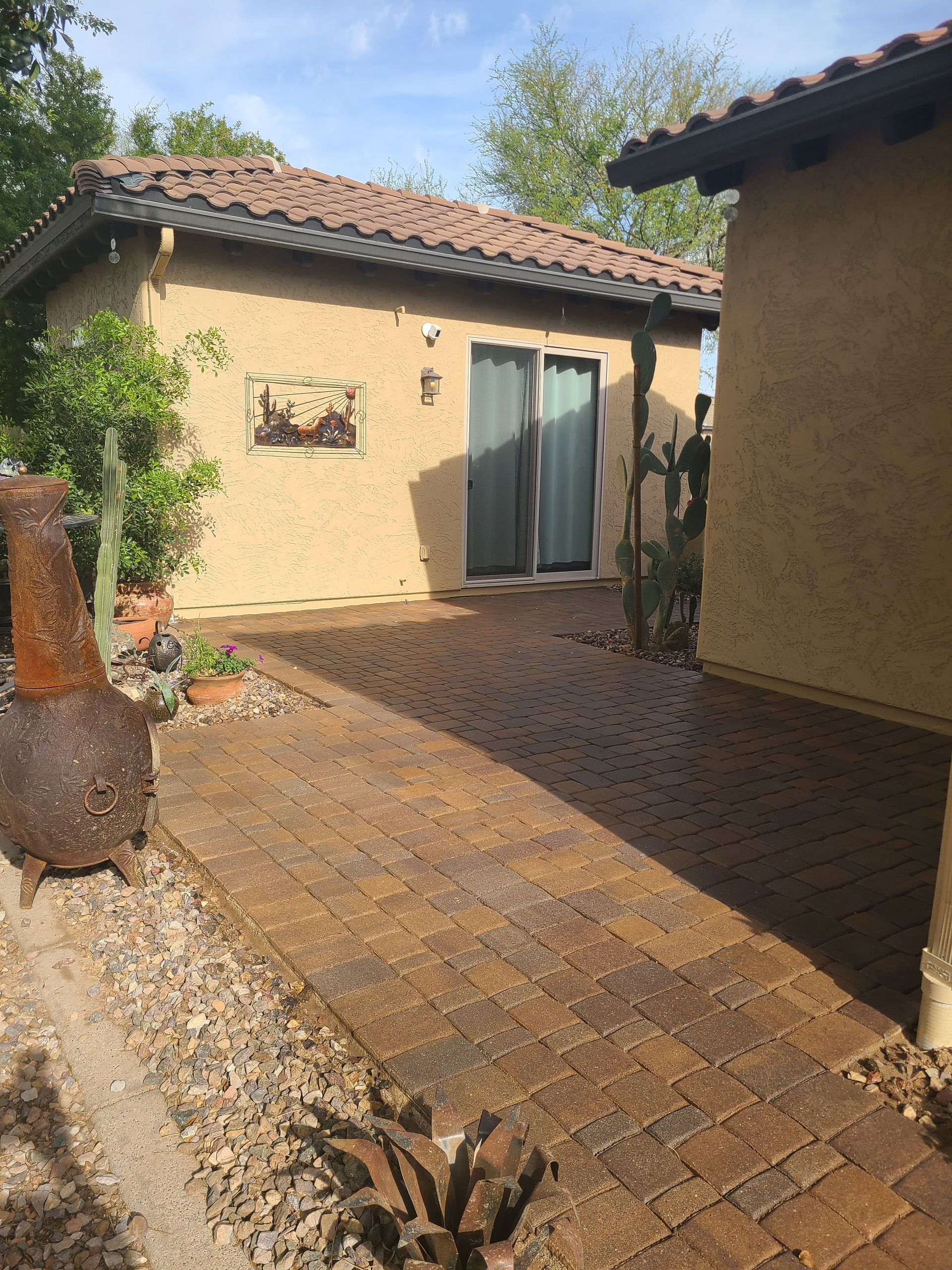 Brick patio leading to a stucco building with a sliding glass door and desert landscaping; rusty fire pit.