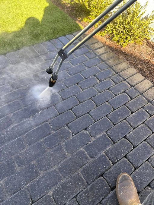 Pressure washer spraying a dark brick patio; boots visible in the foreground, grass in the background.