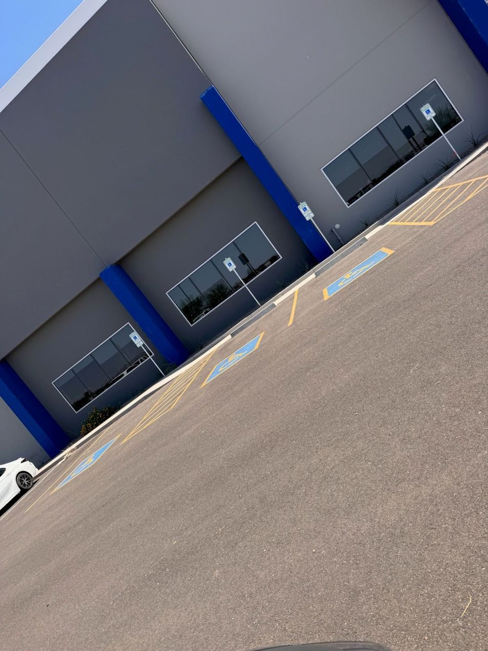 Exterior view of a grey building with blue pillars and windows. Parking lot with disabled parking spaces in front.
