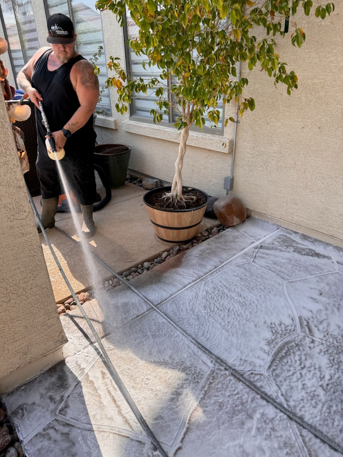Man power washing a concrete patio near a potted tree.