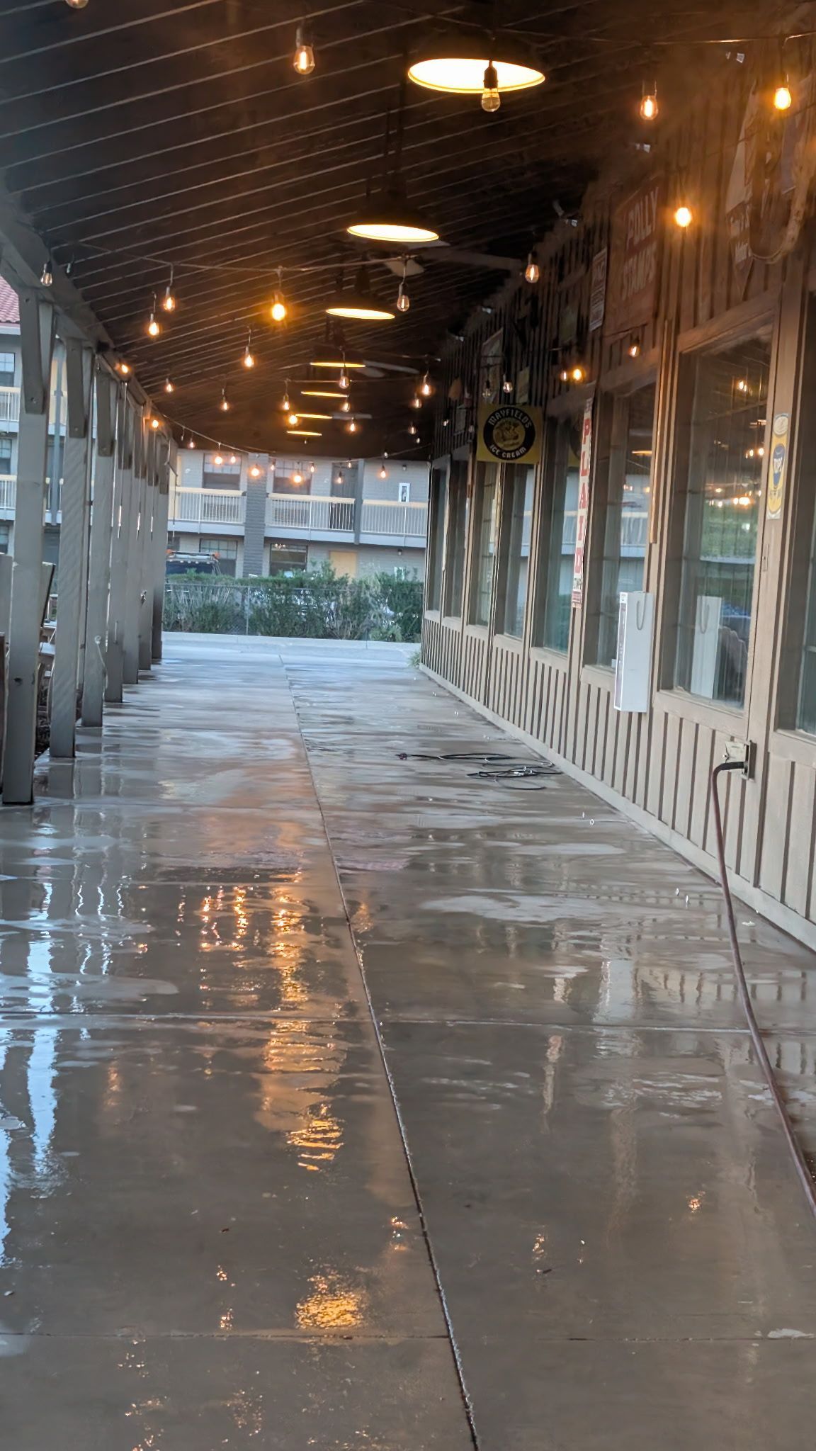 Exterior walkway with wet gray flooring, string lights, and windows. Buildings in the background.