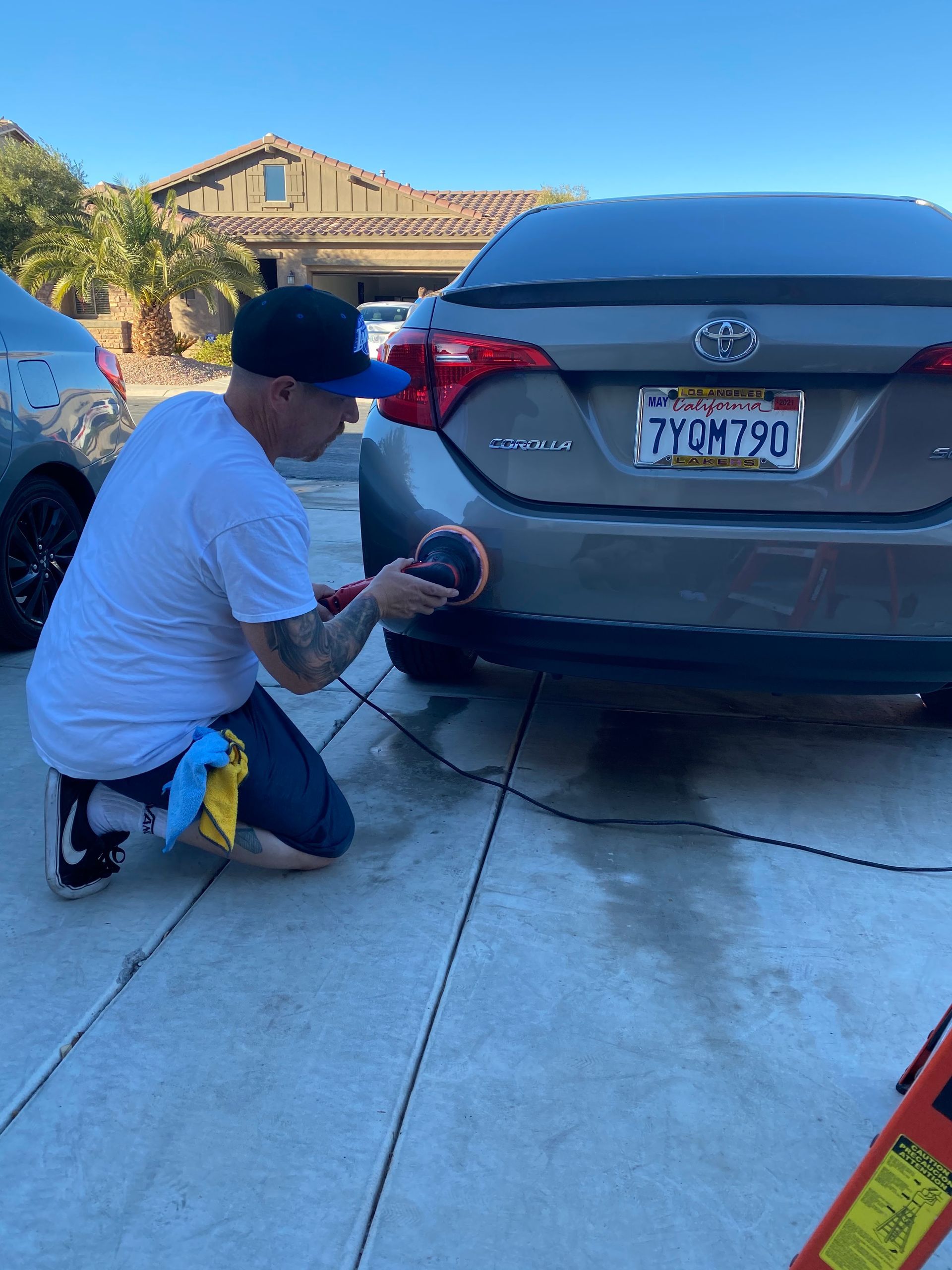 Man polishing a gray Toyota Corolla. Outside on concrete. Holding polisher, kneeling.
