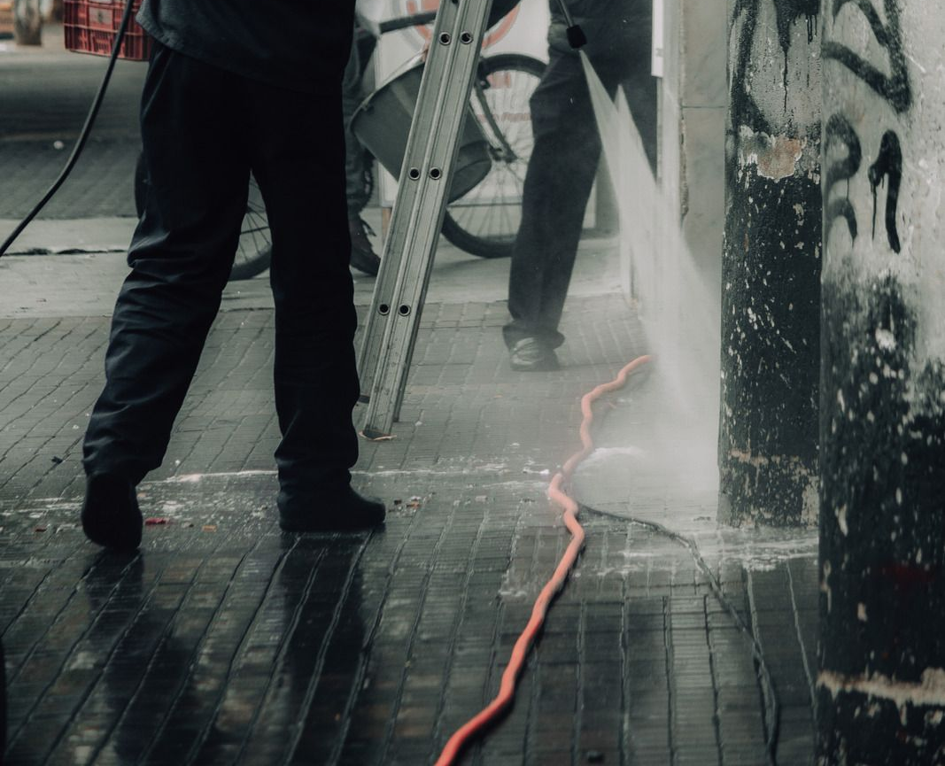 People power washing a wall and sidewalk with high-pressure water, removing graffiti.