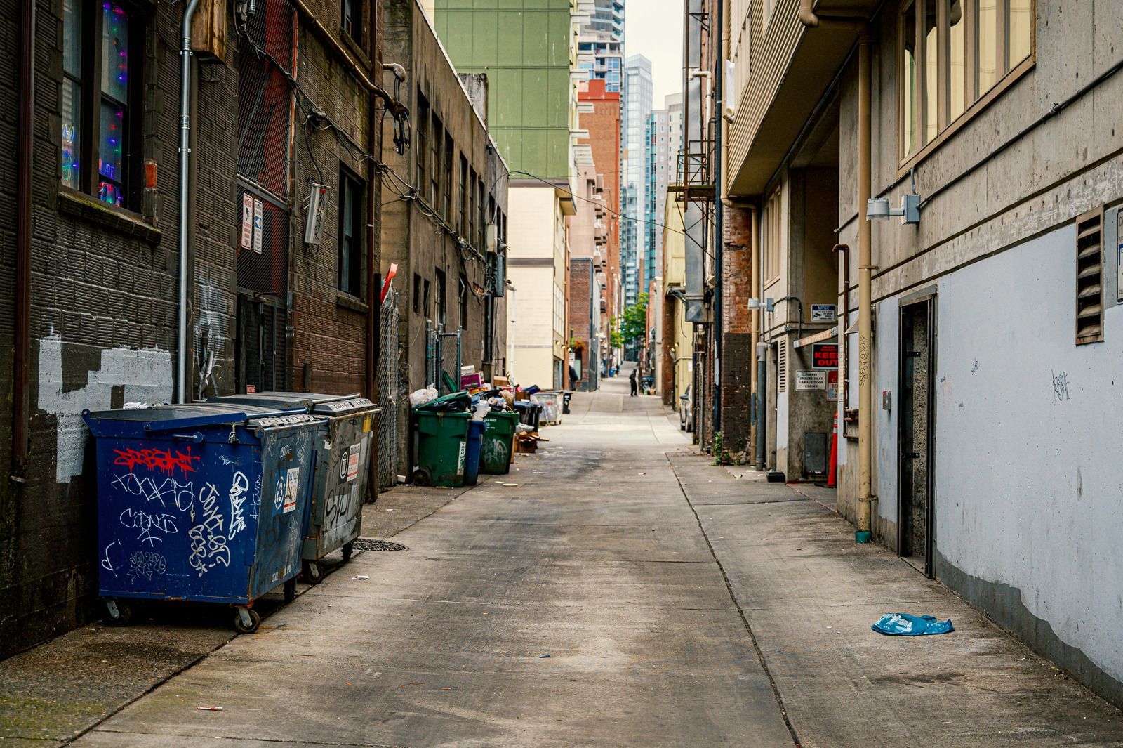 Narrow urban alley with trash bins, aged buildings, and a distant view of skyscrapers.