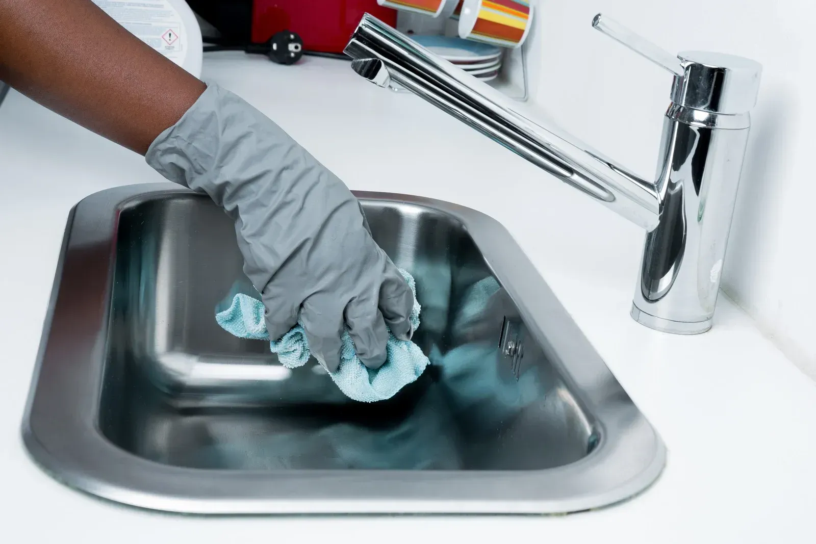 Person with gloved hand wiping a stainless steel sink with a blue cloth.