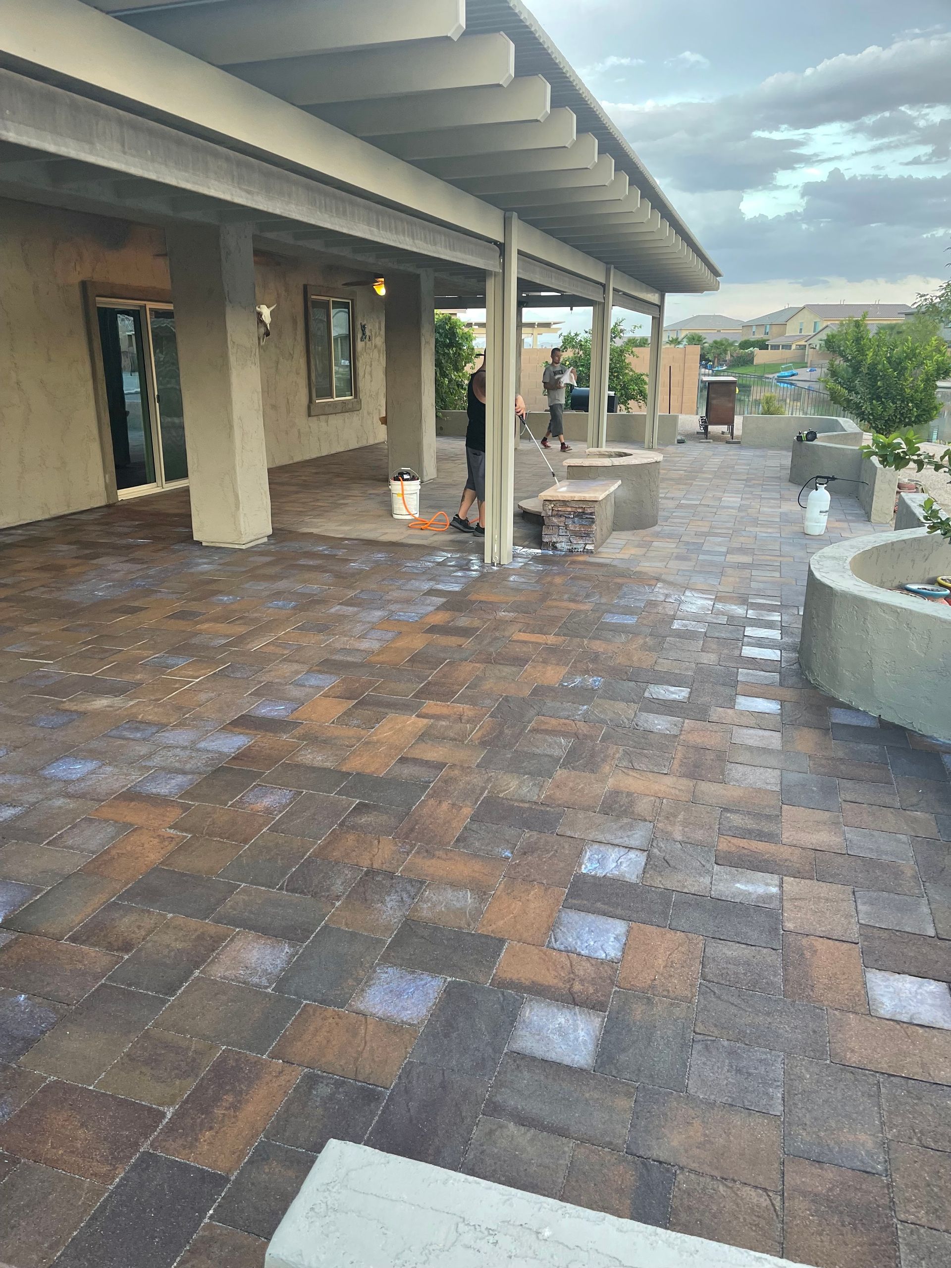 Patio with multicolored brick pavers, covered by a light-colored pergola. Workers are visible; a natural, outdoor setting.