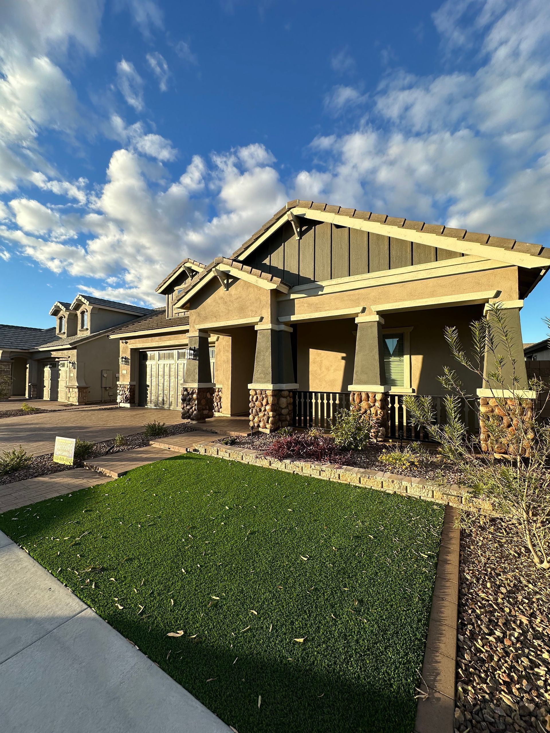 House with tan and gray exterior, green lawn, blue sky, and clouds.