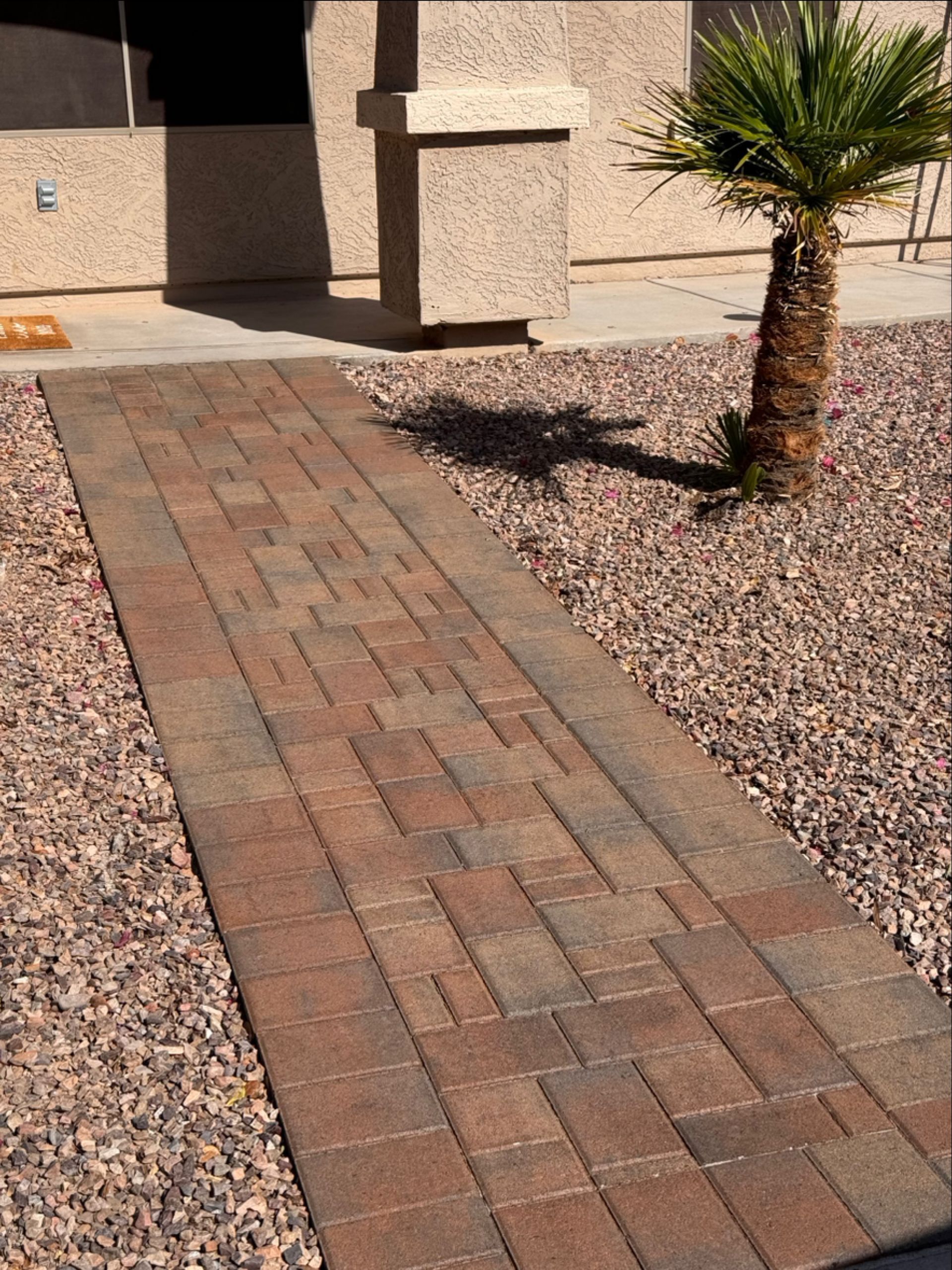 Brick walkway leading to a beige building entrance, bordered by gravel and a small palm tree.