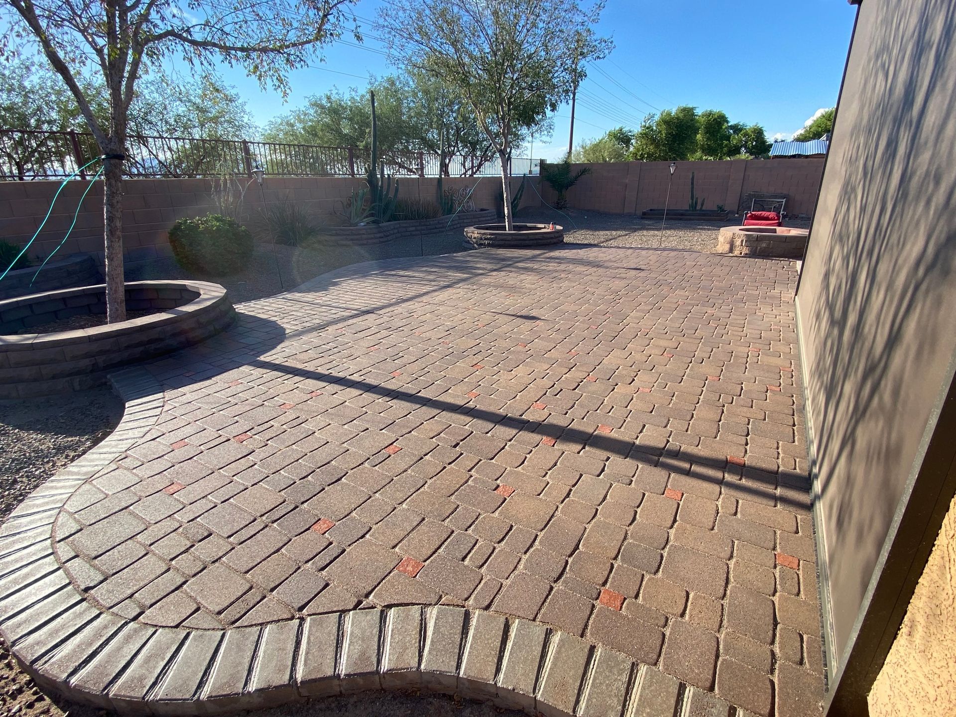 Brick patio with curved edges, trees, and a low wall. Sunny outdoor setting.