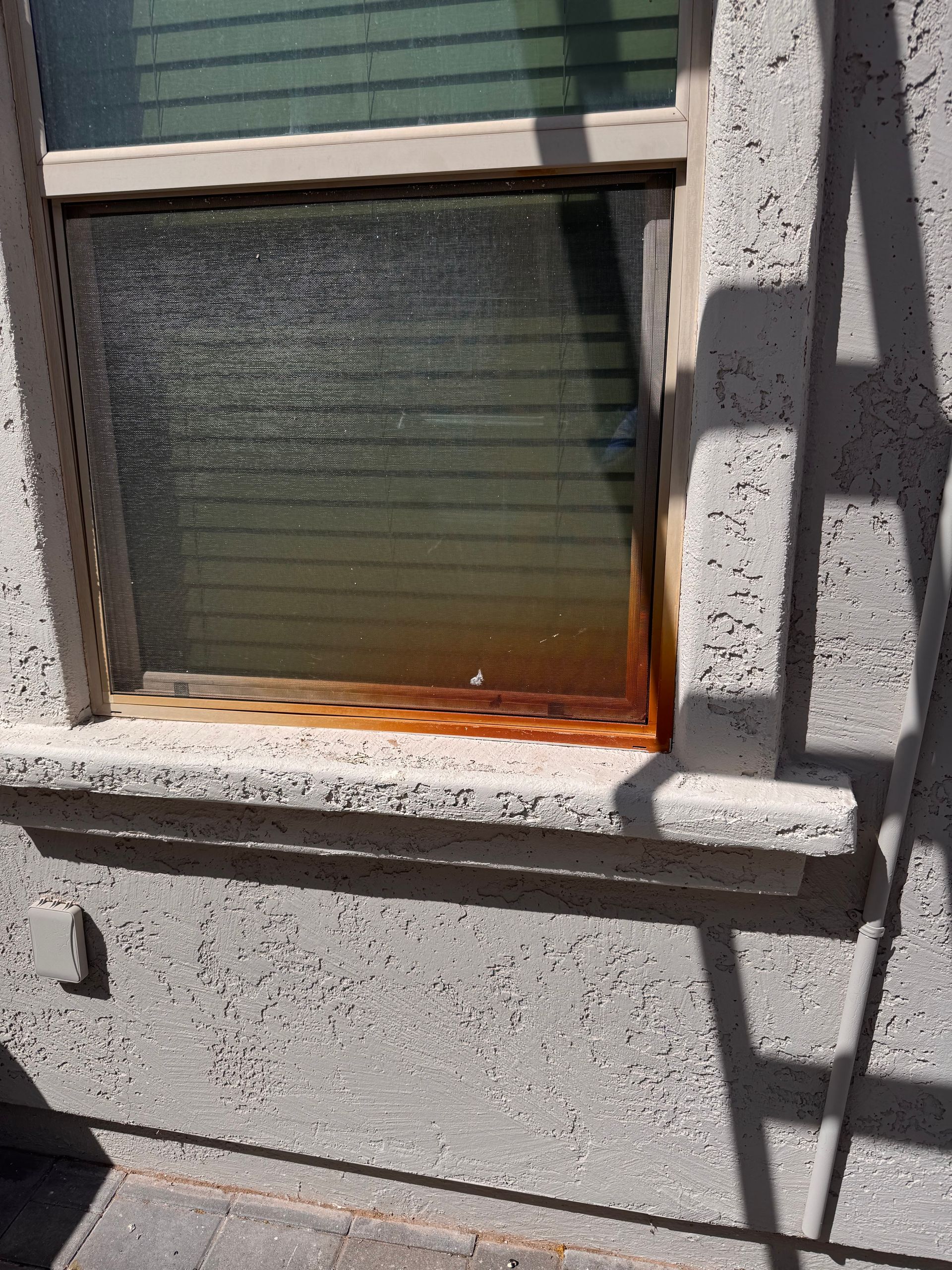 Window with rusted metal frame and screen, set in textured stucco wall, lit by sunlight.