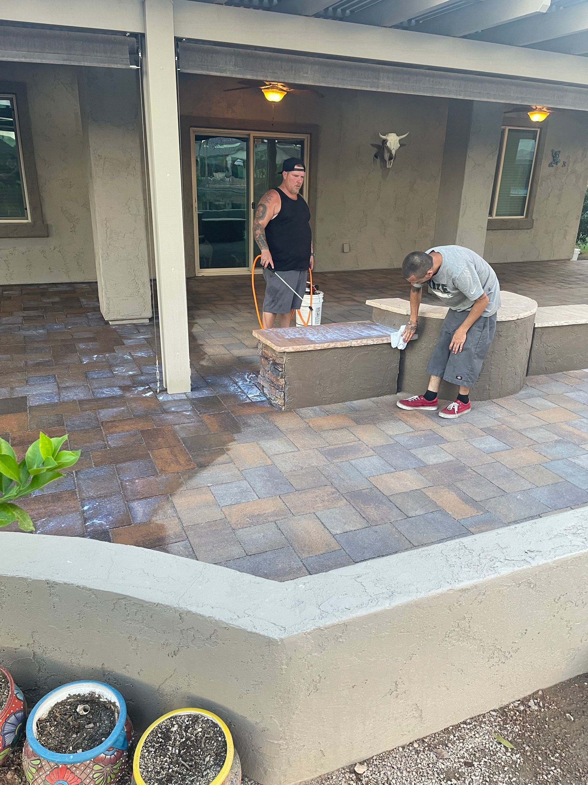 Two people working on a brick patio. One hoses down the bricks, the other scrubs a low wall.