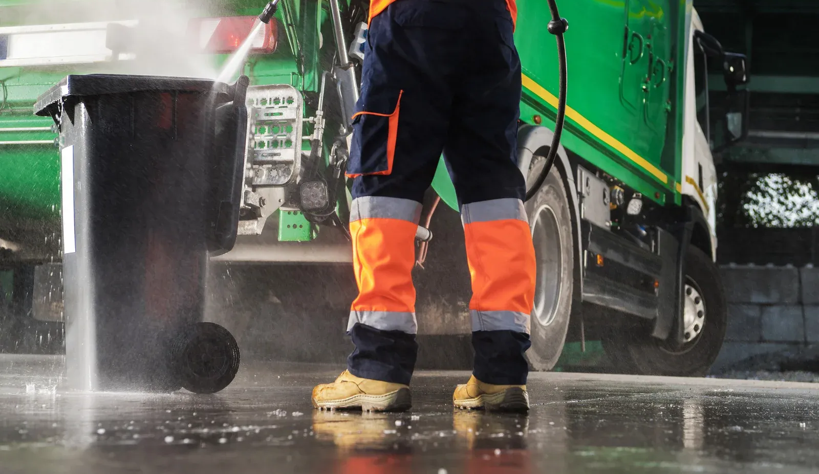 Worker in orange and navy work clothes power-washes a black trash bin near a green garbage truck.