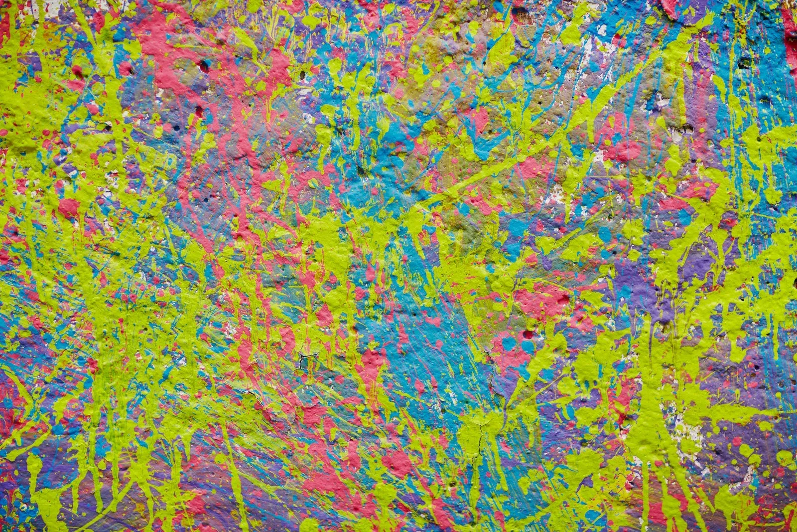 Colorful paint splatters on a textured wall. Primarily pink, blue, green, and yellow.