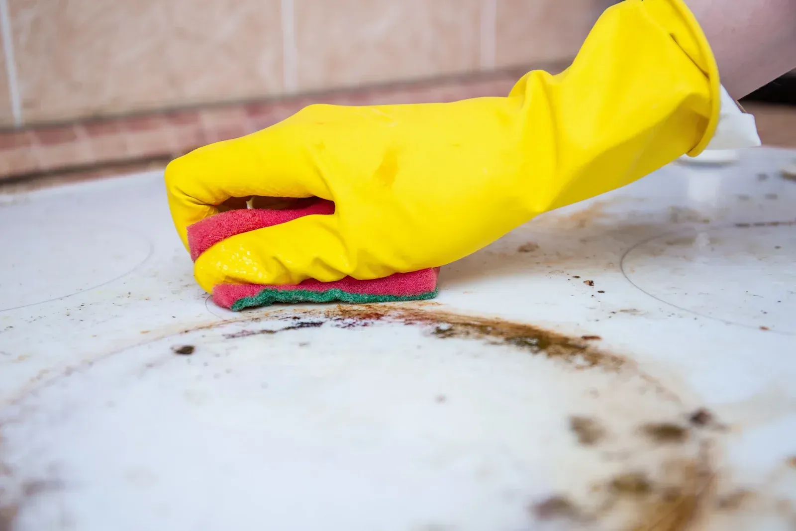 Yellow-gloved hand scrubbing a dirty white stovetop with a red and green sponge.