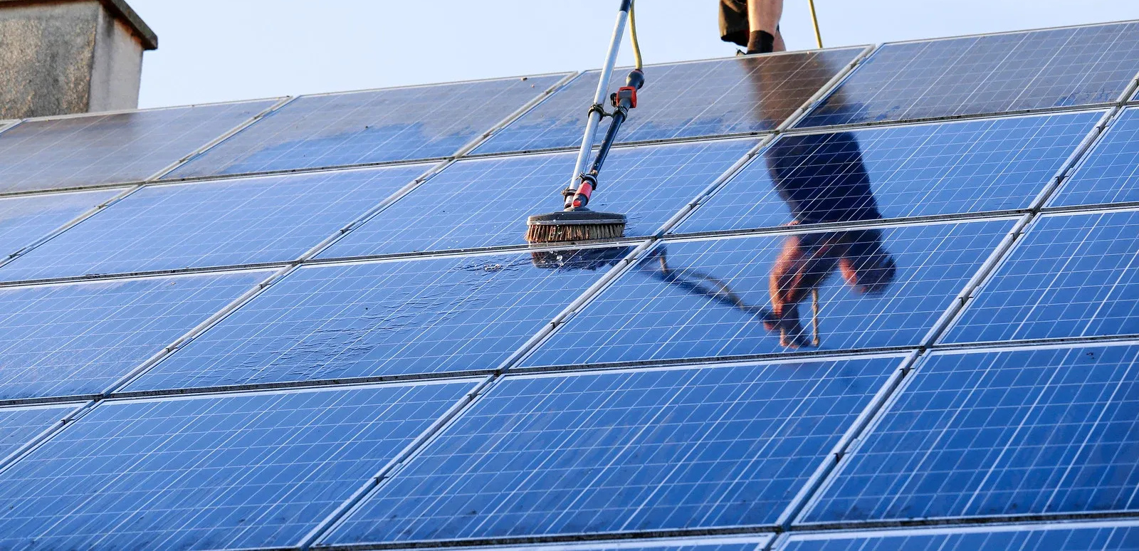 Person cleaning solar panels on a roof with a long-handled brush; the panels are blue.
