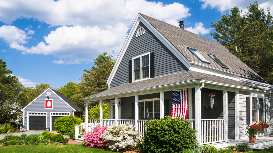 Blue house with white porch, American flag, garage, and garden under a blue sky.