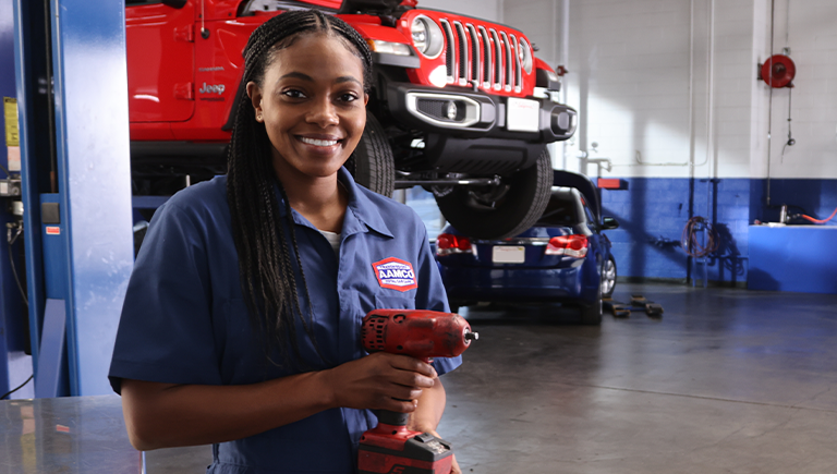 AAMCO mechanic holding a tool, smiling at the camera in a garage with cars on lifts.