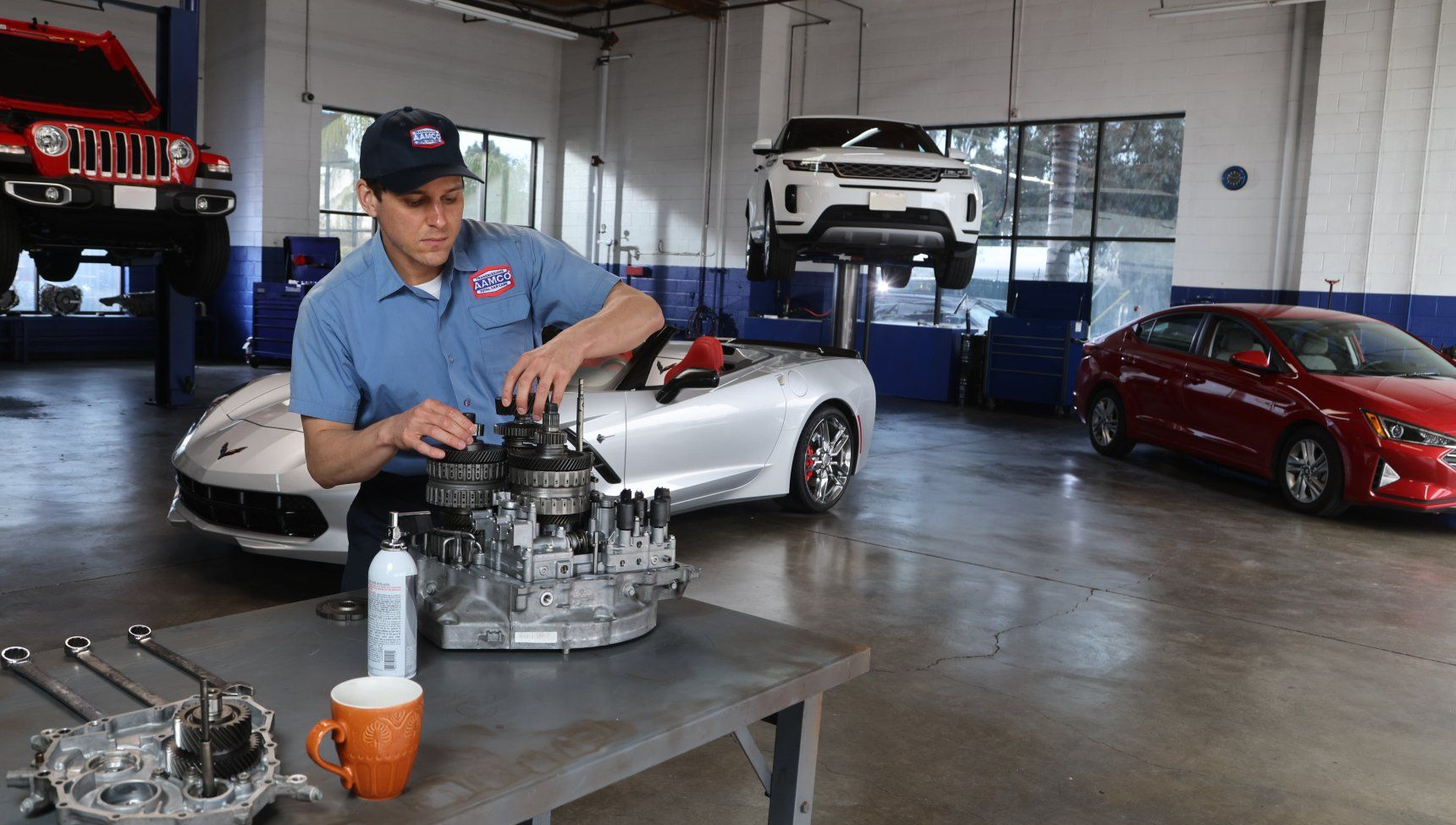 AAMCO mechanic working on car transmission in auto shop.  Silver car and red car are in the background, another on lift.