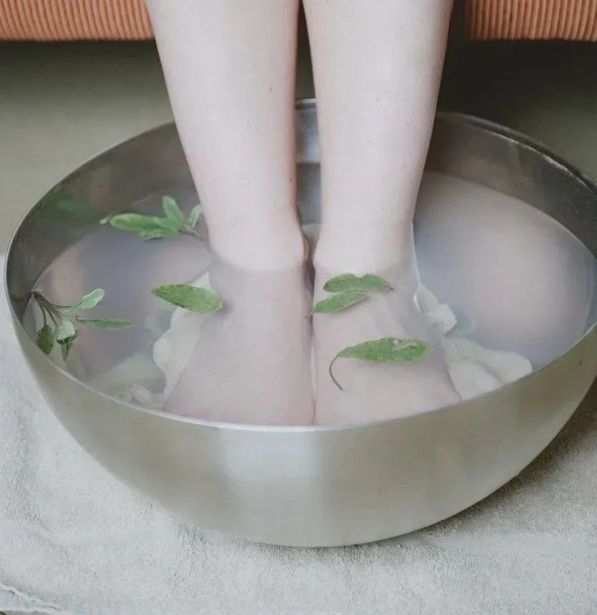 Feet soaking in a metal bowl of cloudy milk with green leaves, suggesting a spa foot bath.