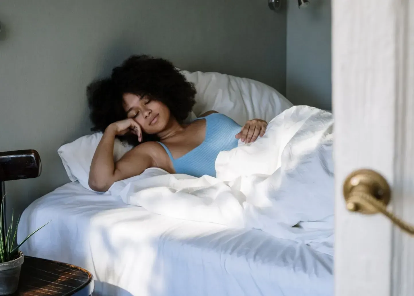 Woman sleeping in a white bed beside a window, with blue bedding and a hand resting on her face