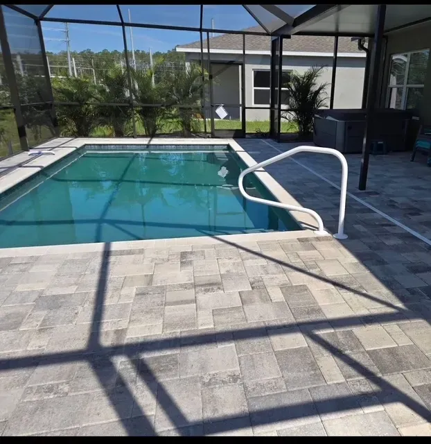 Swimming pool with blue water, surrounded by gray brick patio enclosed by a screened structure.
