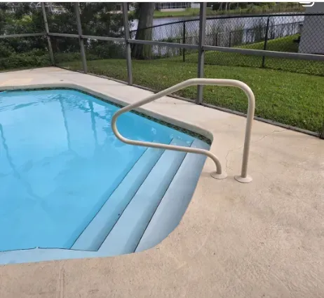 Pool with built-in steps, a beige handrail, and a concrete deck, set beside a fence and green lawn.