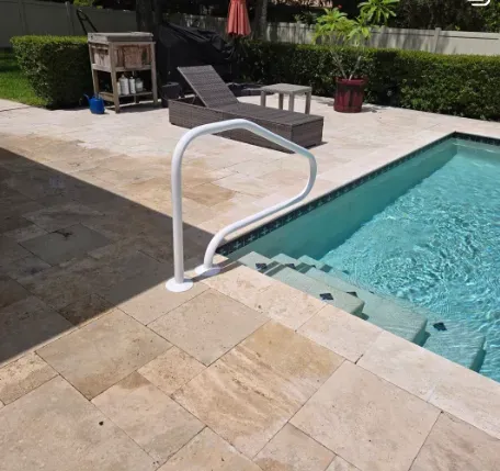 White pool handrail by a blue pool, stairs, tan patio, lounge chair.