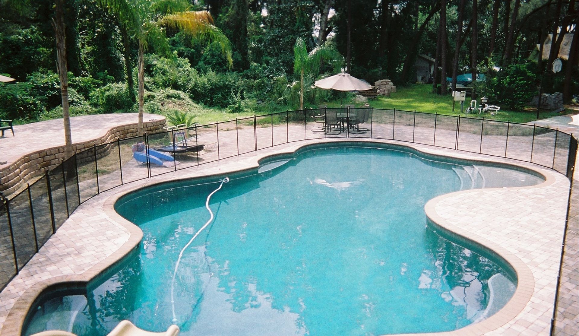 A kidney-shaped swimming pool with a surrounding stone patio, surrounded by a black fence. Trees and greenery are in the background.