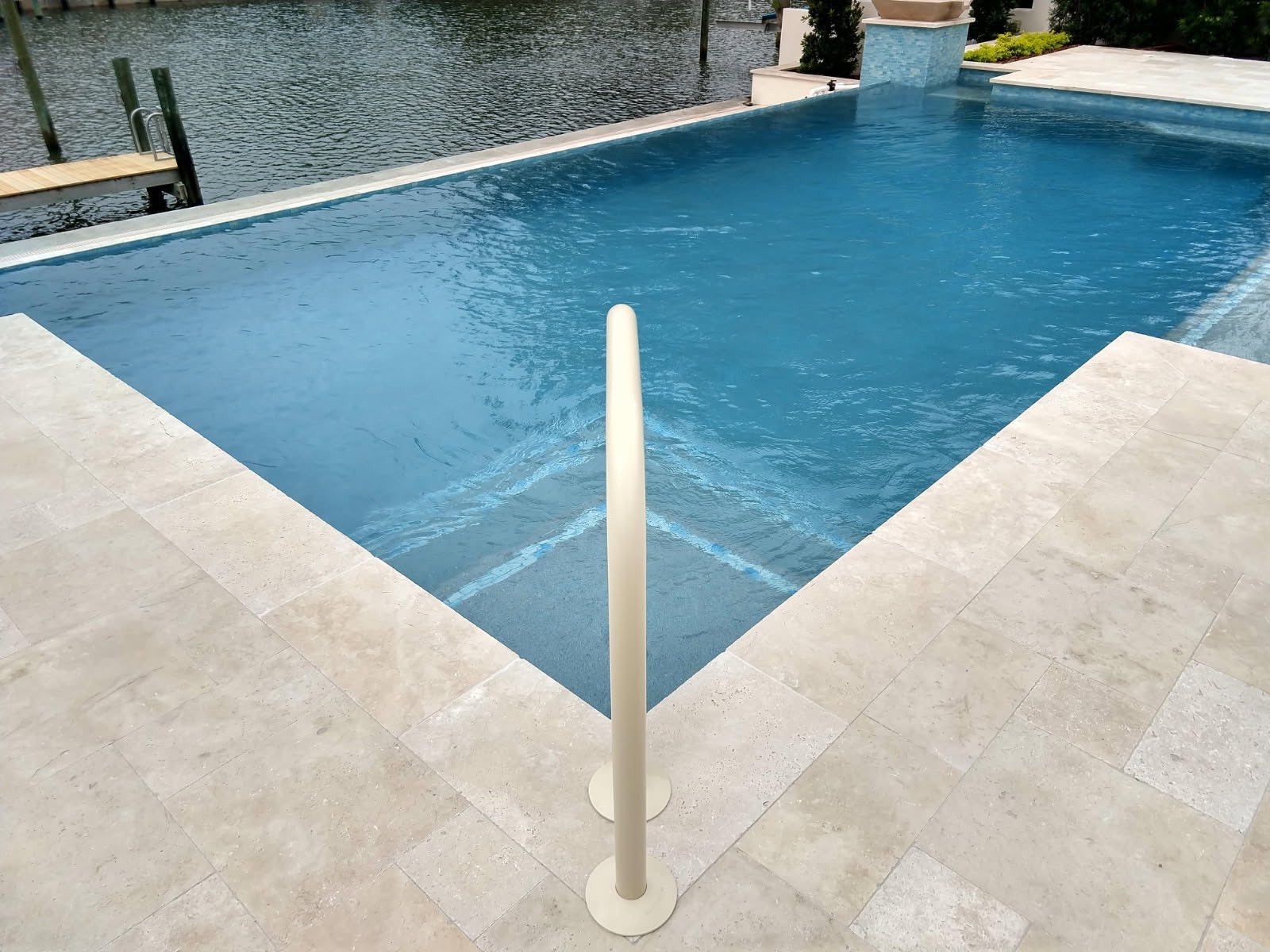 Swimming pool with tan stone deck and a white handrail for entry. Blue water.