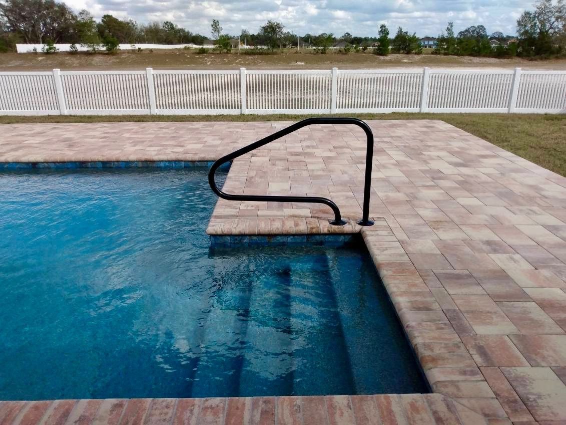 Pool steps with a black handrail, brick coping, and a white picket fence in the background.