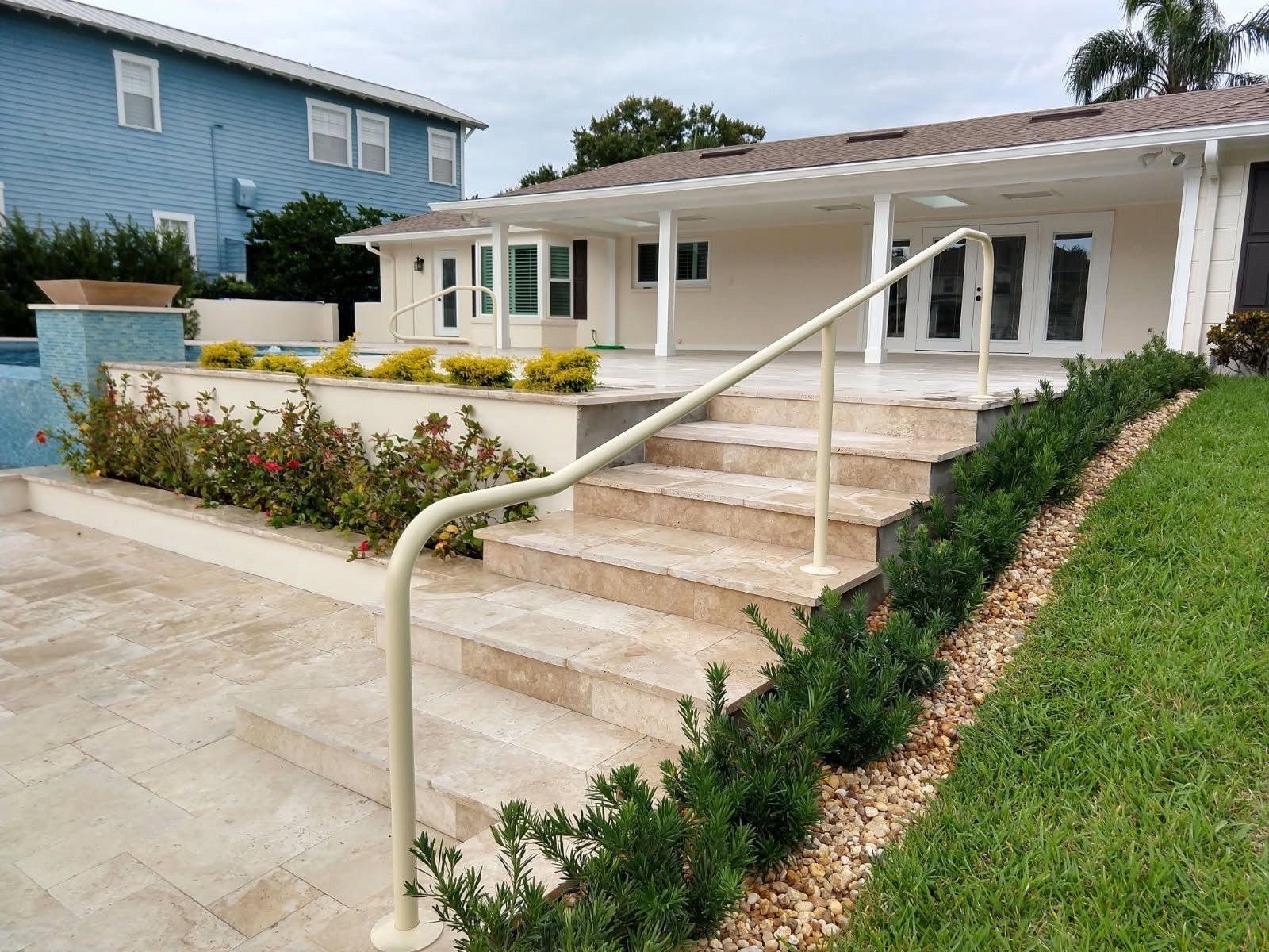 Beige handrail on stone steps leading to a house with a pool and garden; lush landscaping.