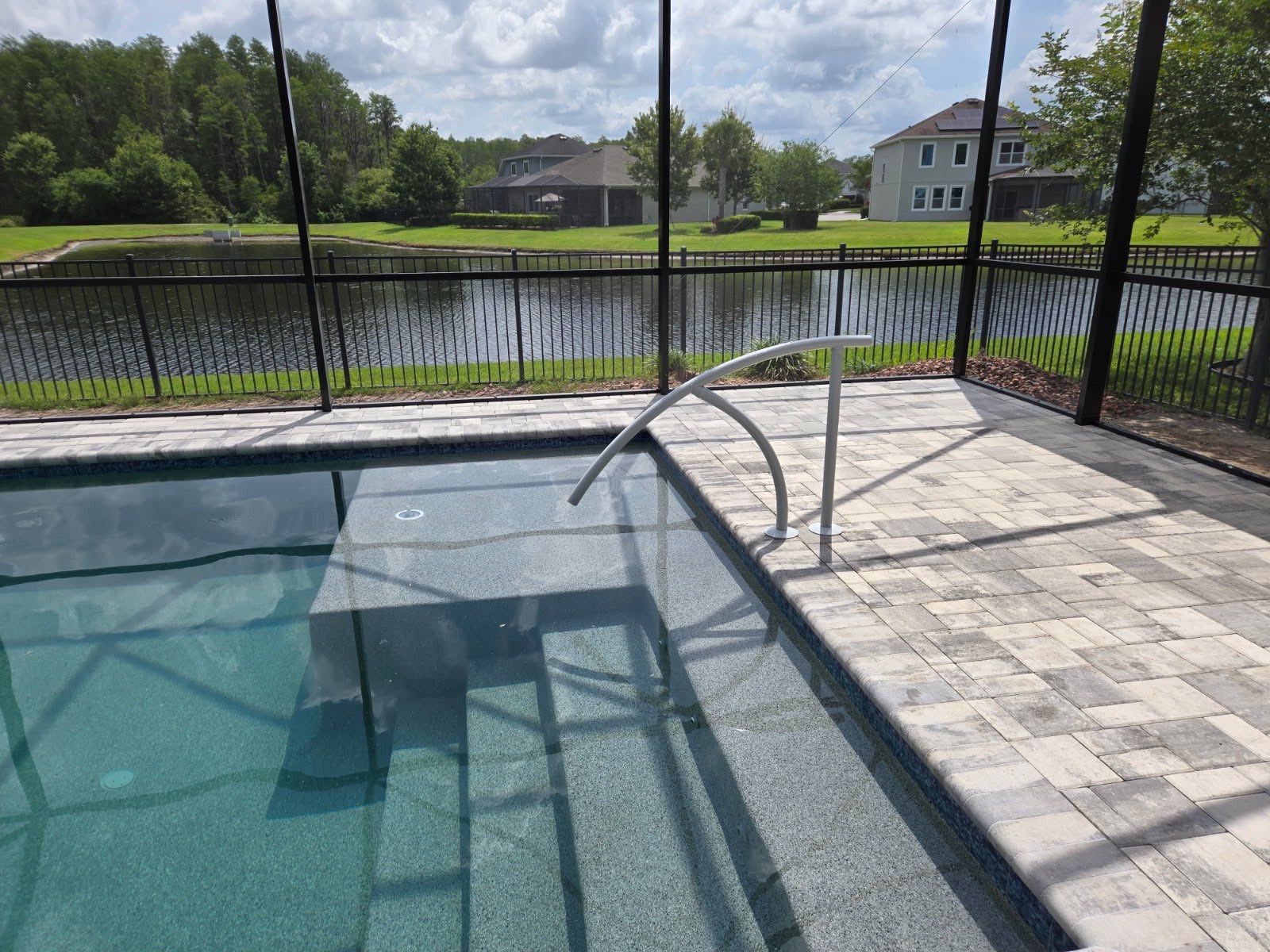 Pool with steps, handrail, and patio, overlooking a pond with a black fence and trees under a cloudy sky.